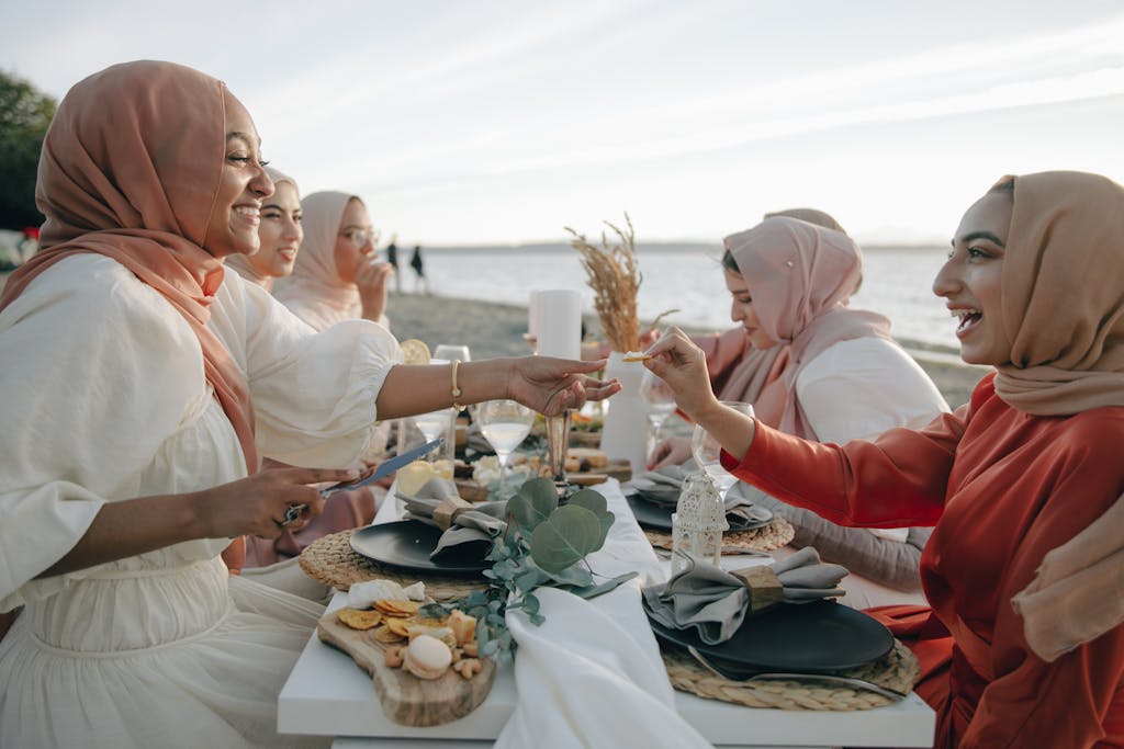 Group of women in hijabs dining by the sea at sunset, sharing laughter and food.